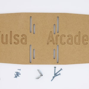 Puede incluir: Un letrero de cartón marrón con el texto "Tulsa Arcades" recortado. El letrero se acompaña de herrajes para el montaje, incluyendo tornillos, arandelas y soportes metálicos.