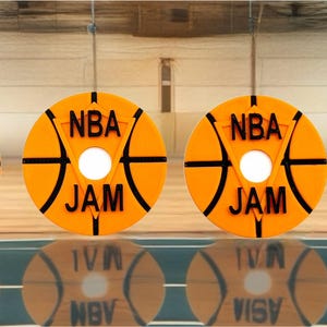 May include: Four orange circular objects with a basketball design and the text "NBA JAM" in black. The objects are arranged in a row on a wooden floor, with their reflections visible below.