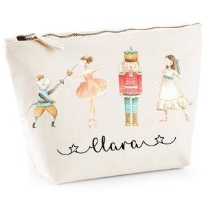 Może przedstawiać: Biała kosmetyczka z zamkiem błyskawicznym w motywie Dziadka do Orzechów. Kosmetyczka przedstawia baletnicę, żołnierza Dziadka do Orzechów, mysz i dziewczynkę w białej sukience. Kosmetyczka jest spersonalizowana imieniem "Clara" czarnym, ozdobnym pismem.