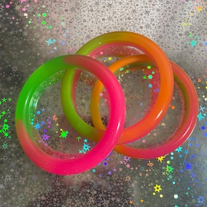 Op de afbeelding: Drie kleurrijke, doorschijnende armbanden in neonroze, groen en oranje. De armbanden zijn in een speels overlappend patroon gerangschikt, tegen een glinsterende achtergrond met sterrenpatroon. Deze accessoires zijn perfect om een vleugje kleur aan elke outfit toe te voegen.