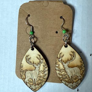 May include: Wooden teardrop earrings featuring a laser-cut design of a deer in a forest. The earrings have silver-colored hooks and a small green bead. The earrings are displayed on a brown earring card.