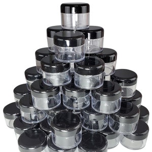 May include: A pile of 20 clear plastic jars with black lids. The jars are stacked in a pyramid shape.