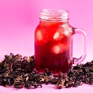 May include: A glass jar filled with a red drink and ice cubes, set against a pink backdrop. Dried, dark red hibiscus flowers surround the jar, suggesting a refreshing drink. The jar has a handle, perfect for a summer beverage.