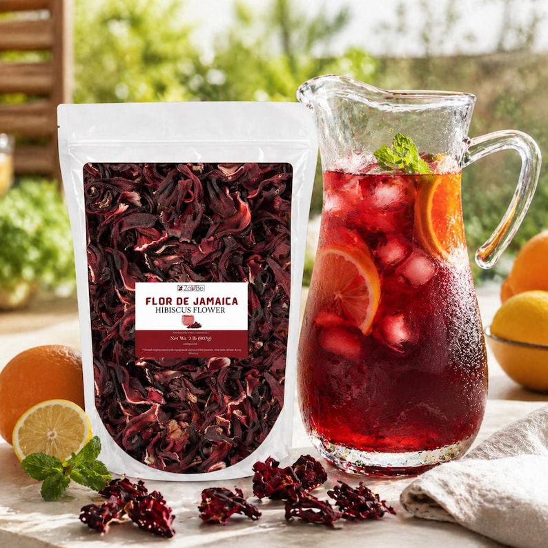 May include: A white bag of dried hibiscus flowers labelled "Flor de Jamaica Hibiscus Flower" beside a glass jug of red hibiscus tea with orange slices and ice. Fresh mint and citrus fruits are also visible.