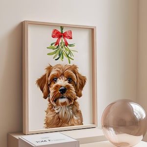 May include: A framed watercolor print of a brown and white dog with a sprig of mistletoe and a red bow above its head. The dog has a fluffy coat and is looking directly at the viewer. The print is in a light wood frame.