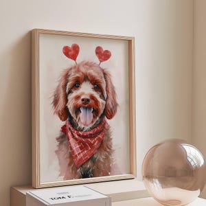 May include: Watercolor painting of a brown dog with red heart-shaped decorations on its head and a red plaid bandana. The dog is framed in a light wood frame, with a neutral background. A decorative glass sphere is in the foreground.