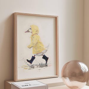 Könnte beinhalten: Eine gerahmte Illustration eines weißen Entchens, das einen gelben Regenmantel und schwarze Gummistiefel trägt und durch eine Pfütze geht.