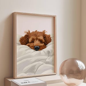 Peut inclure: Une impression encadrée représentant un chien endormi, avec une fourrure brune et un nez noir, reposant sur une couverture blanche. L'œuvre est dans un cadre en bois, sur un fond rose pâle. Une sphère décorative en verre est au premier plan.