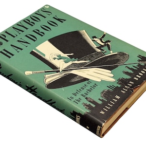 May include: A vintage copy of "The Playboy's Handbook" with a teal cover. The cover illustration includes a black top hat, a woman, and a man in a tuxedo. The title is in large, black letters. The book is angled, revealing its aged, yellowed edges.
