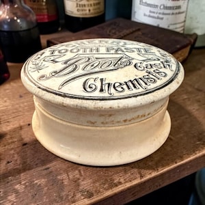 Pode incluir: Pote de cerâmica creme com tampa com a inscrição "TOOTH PASTE Boots Cash Chemists" em escrita preta. O pote tem uma base e tampa arredondadas, com uma aparência ligeiramente envelhecida. O tema é o de um boticário vintage.