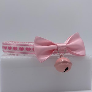 May include: Pink choker with a pink satin bow and a pink bell charm. The choker has a white background with pink hearts.