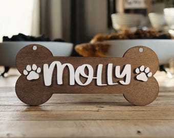 Könnte beinhalten: Ein hölzerner Hundeknochen mit dem Namen "Molly" in weißen Buchstaben und zwei weißen Pfotenabdrücken auf jeder Seite.