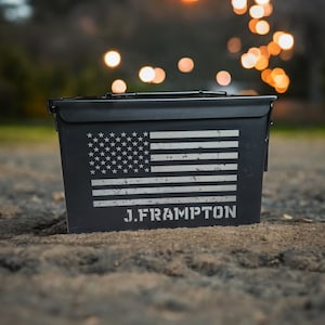 May include: A black metal ammo can with a distressed American flag design and the text "J. FRAMPTON" printed on the front.