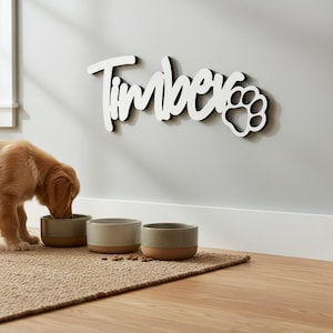 May include: A golden retriever puppy eats from a ceramic bowl on a woven mat. Above, a white wooden sign with the name "Timber" in cursive and a paw print is mounted on the wall. The bowls are neutral colors.