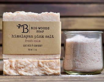 Può includere: Due saponette al sale rosa dell'Himalaya con un'etichetta marrone che recita "BIG WOODS SOAP himalayan pink salt fresh calm GOAT MILK & RAW HONEY NET WT: 5.5 OZ". Un barattolo di vetro pieno di sale rosa dell'Himalaya è accanto al sapone.
