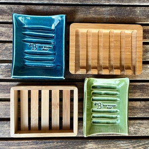 May include: Four soap dishes, two made of wood and two made of ceramic. The ceramic dishes are green and blue and have the words "Big Woods Soap" printed on them. The wooden dishes are made of light brown bamboo.