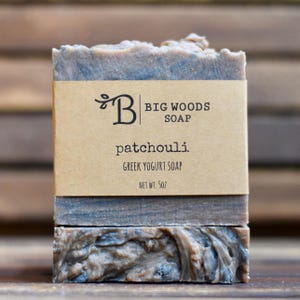 May include: A rectangular bar of Big Woods Soap with a marbled brown and grey texture. A tan paper label wraps around the center, printed with the text Big Woods Soap, patchouli, Greek yogurt soap, and the net weight of 5oz in black ink.