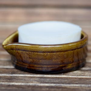 May include: A brown ceramic soap dish with a white bar of soap inside. The dish has a curved design with a small spout on one side. The soap is round and fits snugly within the dish. The dish is on a wooden surface.