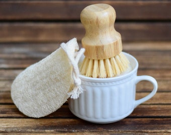 May include: A white ceramic mug with a wooden-handled dish brush and a natural loofah sponge. The brush has light-colored bristles. The mug has a handle and vertical ridges. The loofah is off-white and has a string attached.