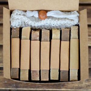 May include: A gift box containing several bars of soap in varying shades of brown and beige, each wrapped in a brown paper sleeve. A white, net-like washcloth and a wooden bead are also included.