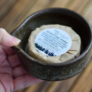 May include: A dark brown ceramic shaving bowl with a spout, holding a wrapped shaving soap. The soap is labeled with ingredients and the text "shaving soap". The label includes a forest graphic. The soap weighs 2 oz.