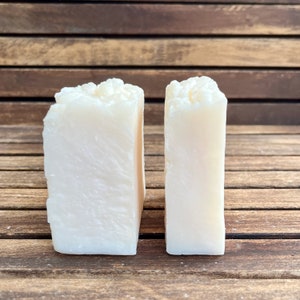 May include: Two rectangular bars of handmade soap. The soap is a creamy off-white color with a textured top. The bars are displayed on a wooden surface.