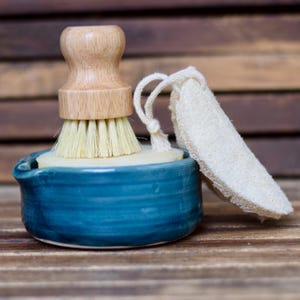 May include: A blue ceramic dish with a wooden brush and a natural loofah sponge. The dish has a small indentation for soap.