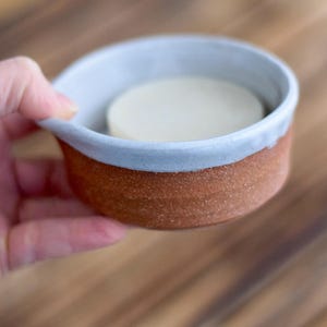 May include: A handmade ceramic soap dish with a light blue rim and a brown base. The dish holds a round, white bar of soap. The dish is held by a hand, and the background is a wooden surface.