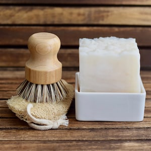May include: A white bar of soap in a white ceramic dish, next to a wooden handled brush with natural bristles and a beige loofah.