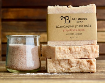 Può includere: Tre saponette di sale rosa dell'Himalaya impilate l'una sull'altra. La saponetta in cima ha un'etichetta di carta marrone con il testo "BIG WOODS SOAP" e "himalayan pink salt grapefruit cream GOAT MILK & RAW HONEY NET WT: 5.5 OZ". Un piccolo barattolo di vetro pieno di sale rosa dell'Himalaya si trova accanto alle saponette.