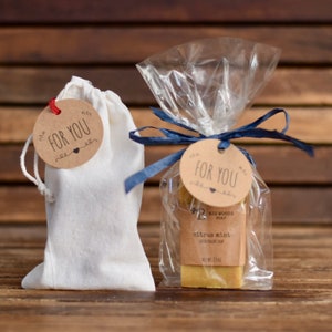 May include: Two gift packages, one in a white muslin bag and the other in a clear plastic bag tied with a blue ribbon. Both packages have a brown paper tag with the text "For You" in black lettering. The plastic bag contains a bar of soap labeled "Citrus Mint" with the brand name "Big Woods Soap".