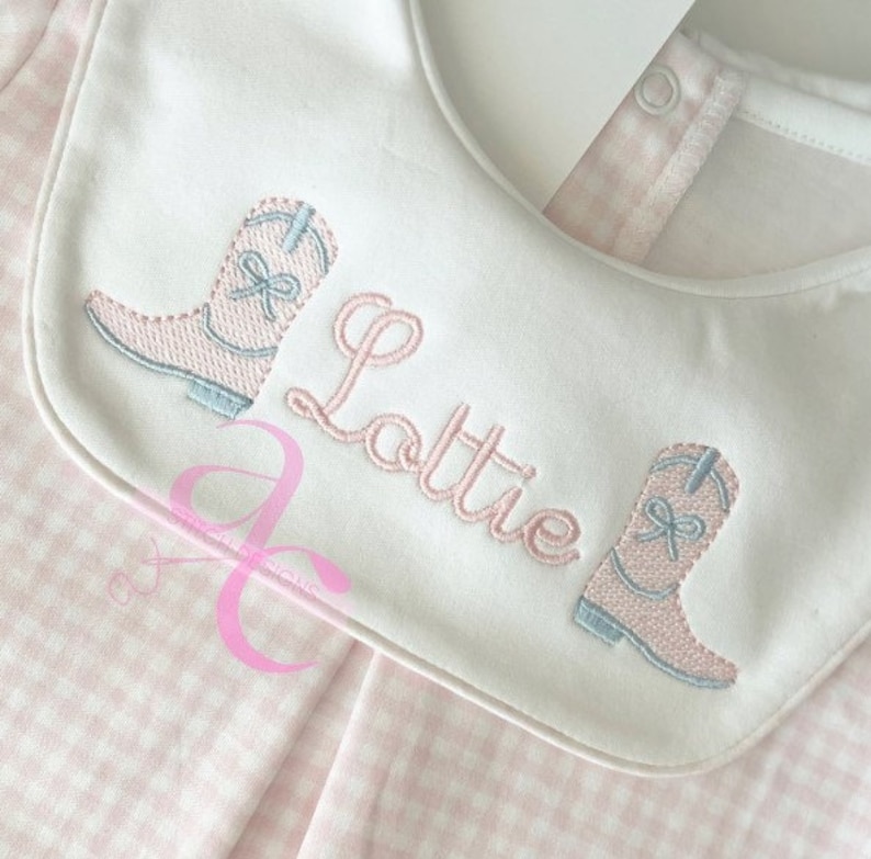 Peut inclure: Bavoir blanc avec motif vichy rose et d&eacute;tails brod&eacute;s. Il pr&eacute;sente deux bottes de cowboy roses avec des accents bleus et le nom "Lottie" en &eacute;criture rose. Le bavoir a une fermeture &agrave; bouton-pression.