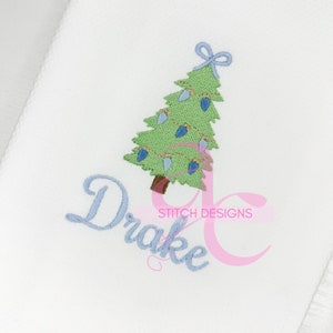 May include: A blue embroidered name "Drake" with a green Christmas tree with lights and a blue bow on top. The text "Stitch Designs" is below the tree.