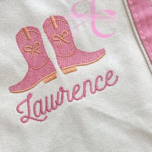 Peut inclure: Un sac fourre-tout en toile avec des bordures roses et des bottes de cowboy roses brod&eacute;es. Le nom "Lawrence" est brod&eacute; en script rose en dessous. Un motif de ruban rose est dans le coin sup&eacute;rieur droit.