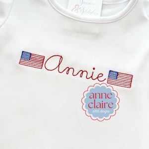 Puede incluir: Camiseta blanca con banderas americanas bordadas en rojo y azul y el nombre "Annie" en cursiva roja. La parte inferior de la camiseta tiene un pequeño círculo azul con el texto "anne claire stitch designs" en blanco.