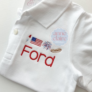 May include: White polo shirt with red embroidered text "Ford" and red, white, and blue embroidered images of a flag, fireworks, and a hot dog. The text "anne claire stitch designs" is embroidered in a pink circle.