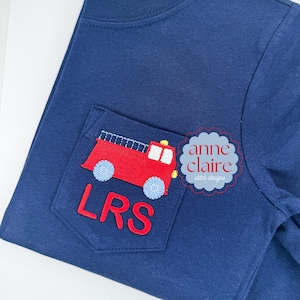 May include: Navy blue t-shirt with a pocket featuring a red embroidered fire truck design. The fire truck has blue wheels and a ladder. The pocket also has the embroidered initials "LRS". The shirt is from "anne claire stitch designs".