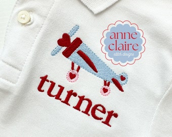 Op de afbeelding: Rood en blauw geborduurd vliegtuig met een hart aan de voorkant, met de naam "turner" eronder geborduurd. Het ontwerp is op een wit poloshirt. Het logo "anne claire stitch designs" staat in een cirkel rechtsboven.
