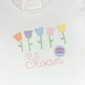 May include: A white onesie with a pink embroidered name "Sloan" and a row of five embroidered tulips in various pastel colors. The tulips are arranged in a line above the name. The onesie is decorated with a small embroidered circle with the text "anne claire stitch designs".