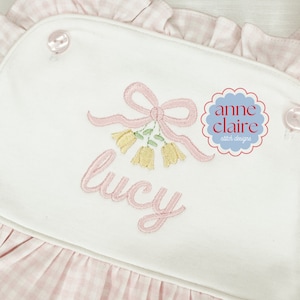 May include: Pink and white embroidered baby dress with a pink bow and yellow flowers, the name "lucy" is embroidered below the bow. The dress has a pink and white gingham pattern on the bottom. The logo "anne claire stitch designs" is in the top right corner.