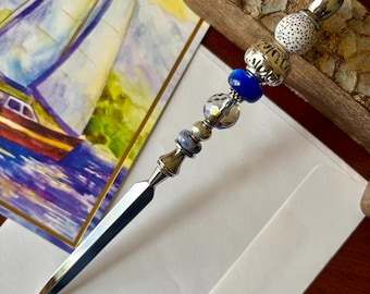 May include: A decorative letter opener with a silver blade and a beaded handle. The handle features blue, white, and clear beads, along with silver accents. A watercolor painting of a sailboat is in the background.