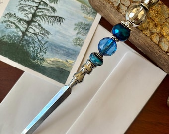 May include: A silver letter opener with a blade and a decorative handle. The handle features blue and clear glass beads. A watercolor painting of a lake scene and white envelopes are in the background.