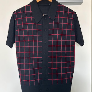 May include: A black short-sleeve knit polo shirt featuring a prominent red grid pattern across the front. The garment includes a classic collar, a central button-down placket with matching black buttons, and ribbed cuffs and hem for a structured fit.