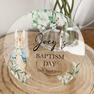 May include: A round, clear acrylic sign with a green floral border and a Peter Rabbit illustration. The sign says "Joey's Baptism Day 14th April 2024".