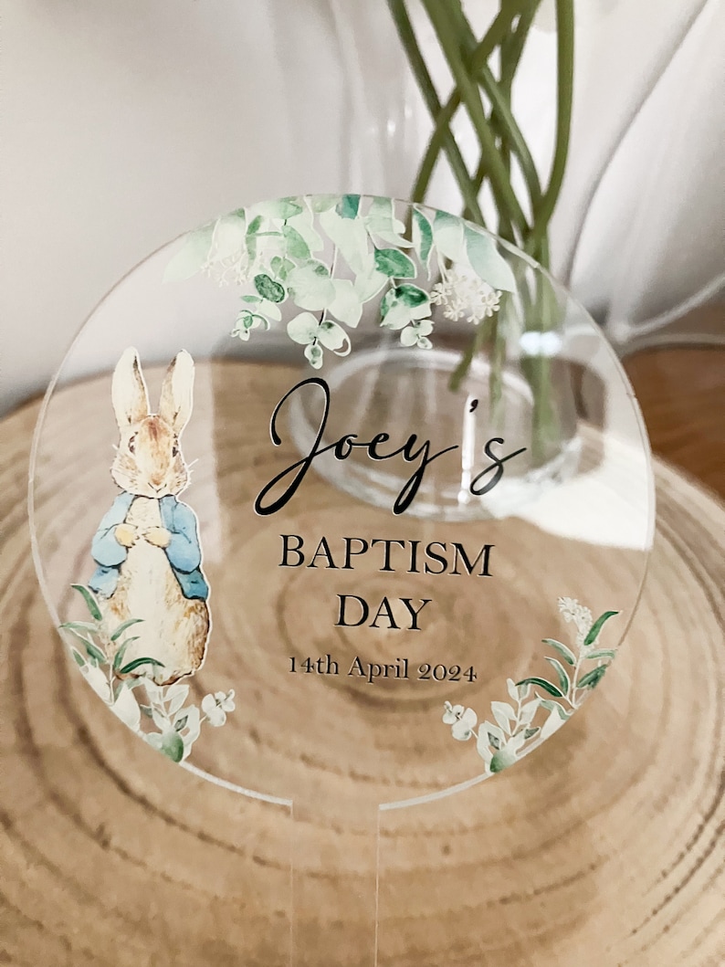 May include: A round, clear acrylic sign with a green floral border and a Peter Rabbit illustration. The sign says "Joey's Baptism Day 14th April 2024".