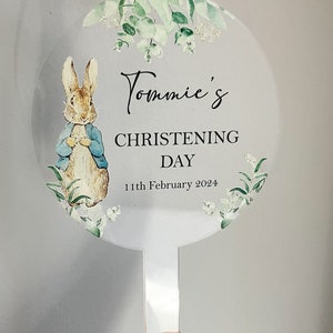 May include: A white round acrylic cake topper with a green floral design and a cartoon image of Peter Rabbit. The text "Tommie's Christening Day 11th February 2024" is printed on the topper.