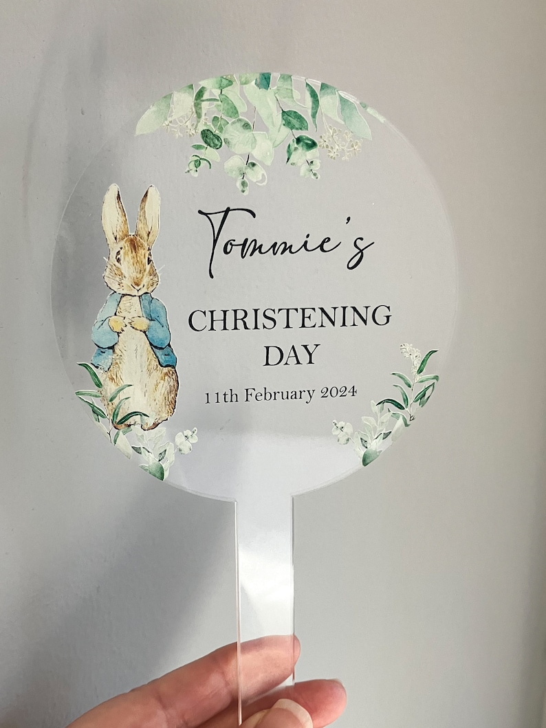 May include: A white round acrylic cake topper with a green floral design and a cartoon image of Peter Rabbit. The text "Tommie's Christening Day 11th February 2024" is printed on the topper.