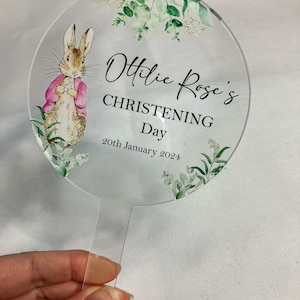 May include: A round, clear acrylic cake topper with a watercolor illustration of a rabbit and greenery. The text "Ottilie Rose's Christening Day 20th January 2024" is printed on the topper.