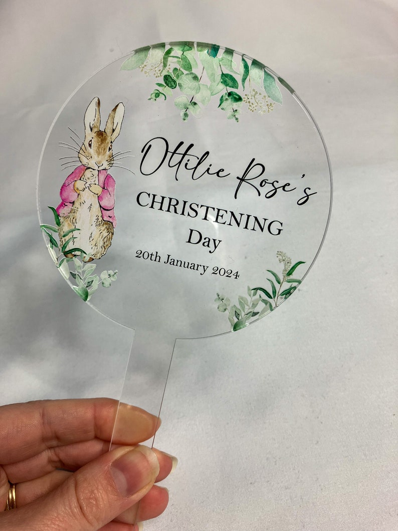 May include: A round, clear acrylic cake topper with a watercolor illustration of a rabbit and greenery. The text "Ottilie Rose's Christening Day 20th January 2024" is printed on the topper.