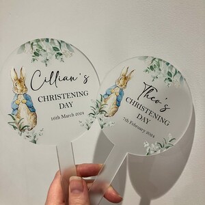 May include: Two round, clear acrylic signs with white floral designs and a watercolor illustration of Peter Rabbit. The signs have the text "Cillian's Christening Day 16th March 2024" and "Theo's Christening Day 7th February 2024".